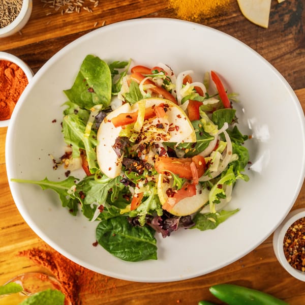 Fresh green salad with pear slices, red peppers, and onions, surrounded by spices on wood.