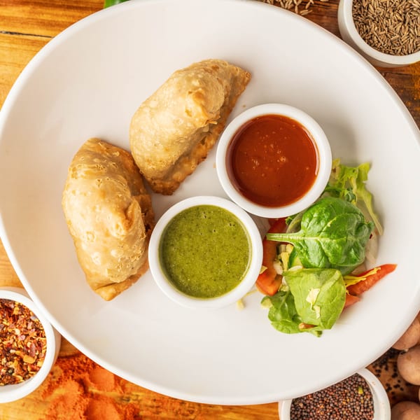 Two golden-brown samosas with green and red chutneys, salad, and spices on a white plate.