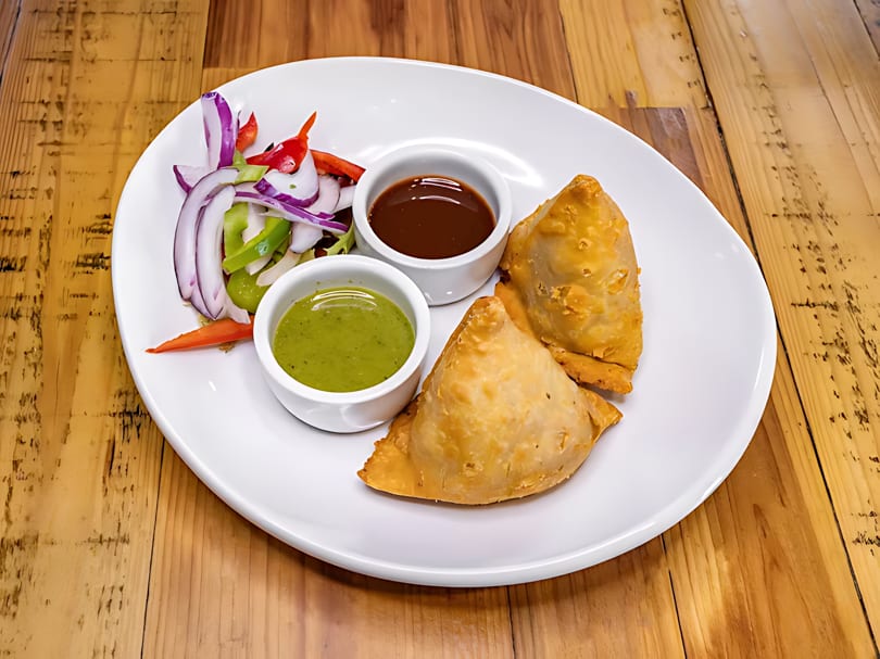 Two golden samosas with green and brown dipping sauces, and a side salad on a white plate.