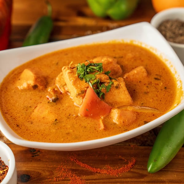 A white bowl of chicken curry with cilantro and tomato, surrounded by spices and vegetables.
