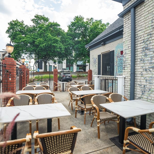 Outdoor patio dining area with marble-top tables and woven chairs next to a brick building.