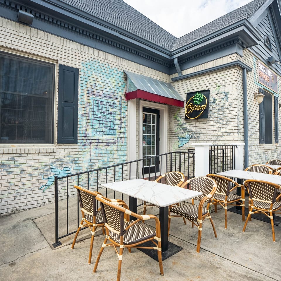 Outdoor patio of Sizzling Spices restaurant with tables, chairs, and a neon "Open" sign.