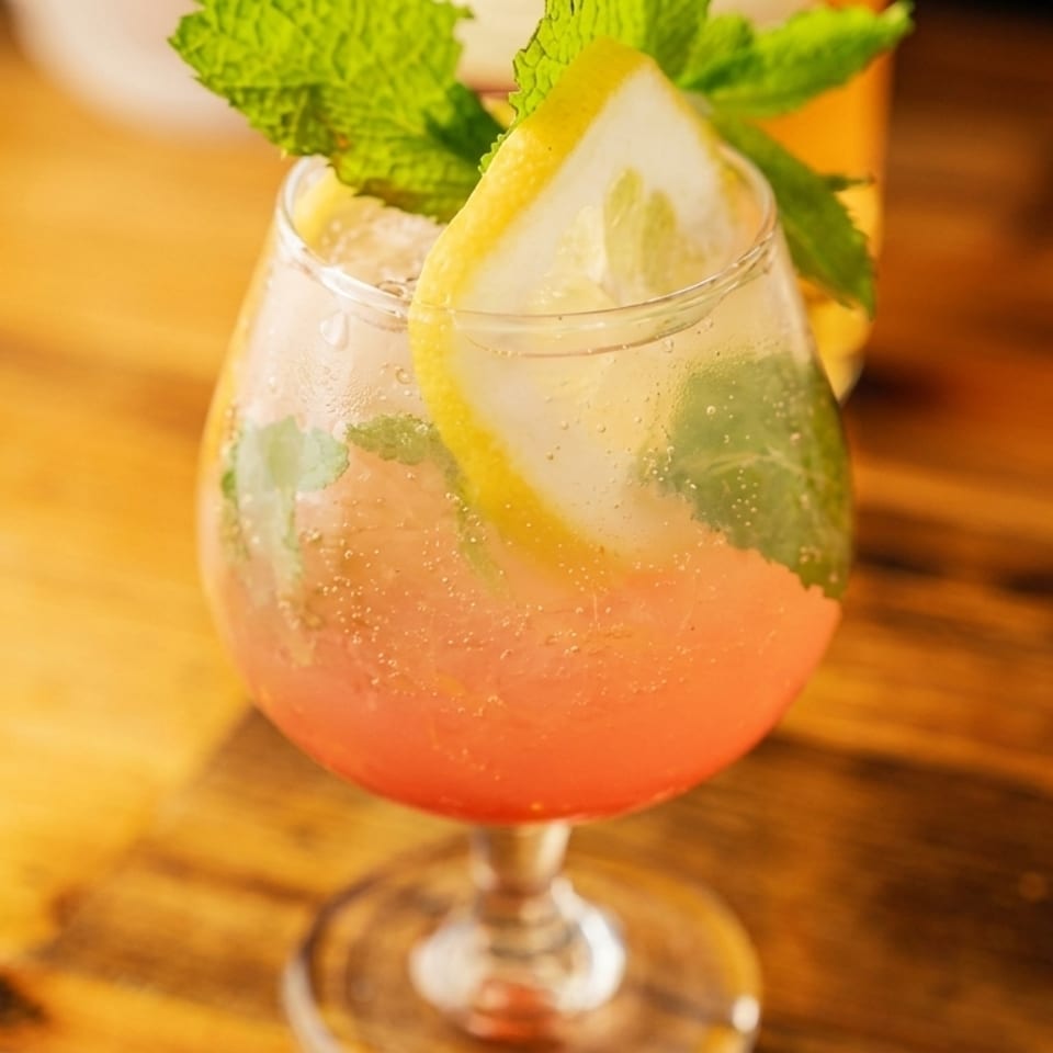 Pink cocktail with lemon and mint garnish in a stemmed glass on a wooden table.