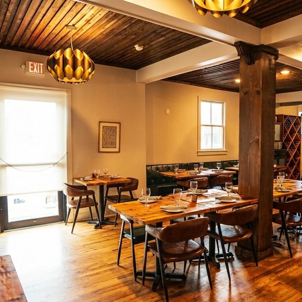 Warmly lit restaurant interior with wooden tables, leather chairs, and a bar area in the background.